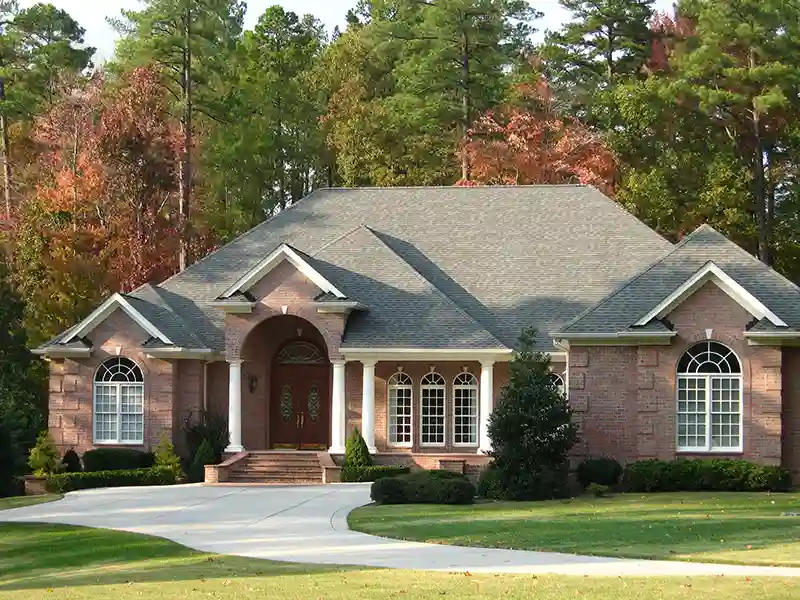 Front view of a higher-end home with gray shingles on its roof and a forest area behind it with tall trees